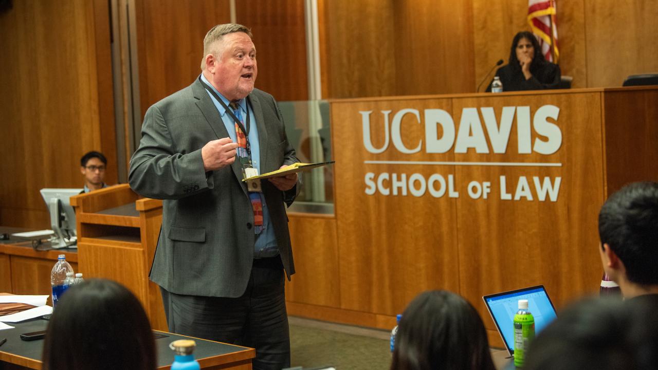 Man in a suit speaking to a group of people. A judge, sitting at their desk is behind the man speaking.