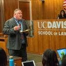 Man in a suit speaking to a group of people. A judge, sitting at their desk is behind the man speaking. 